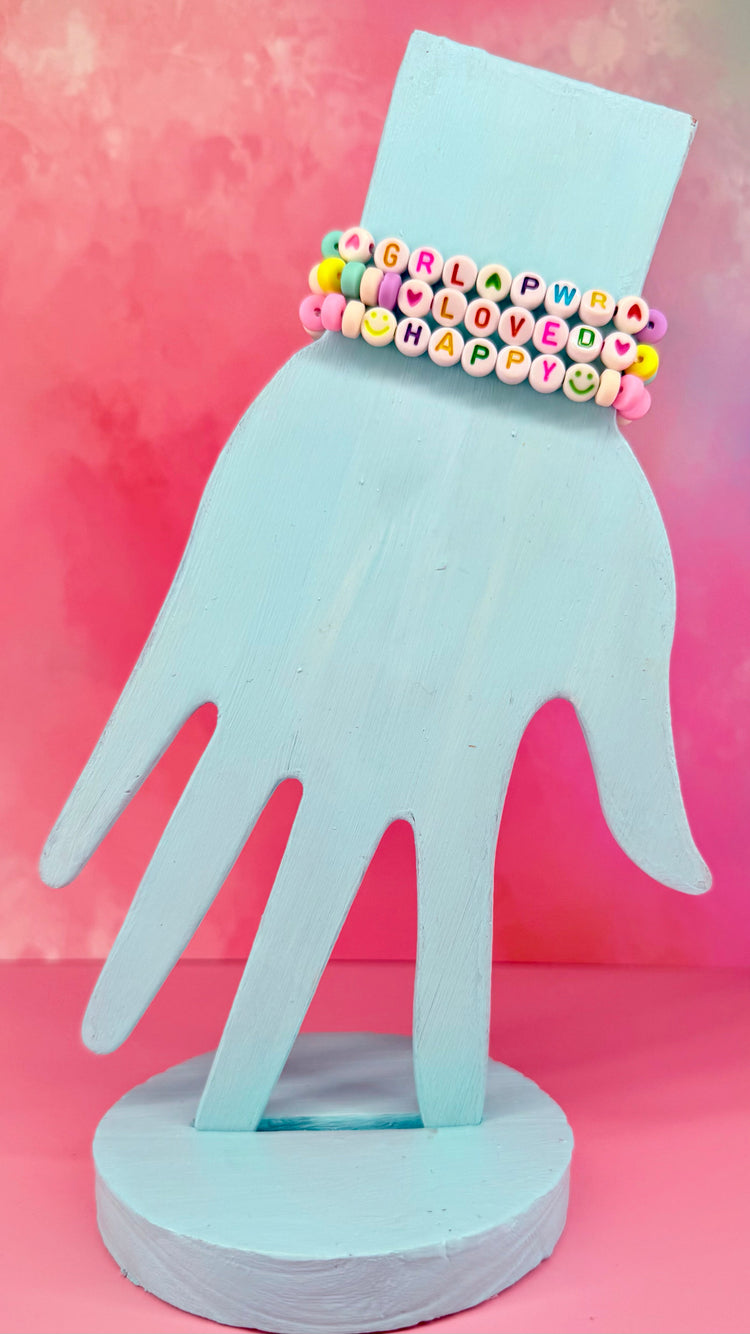 Stacked pastel bracelets with beaded words like GRLPWR, LOVED, and HAPPY on blue display hand
