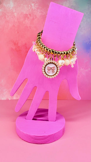 Gold beaded charm bracelet with bow medallion on a pink hand display